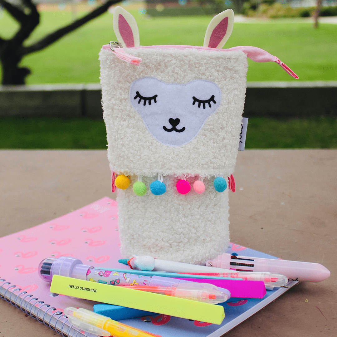 lifestyle image of llama stand up pencil case shown with pink flamingo notebook and assortment of pens